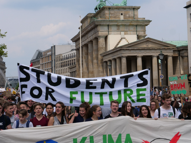 Gruppe von Studenten marschiert in Berlin mit einem leuchtend bunten "Students for Future"-Schild vor einem Hintergrund aus Gebäuden, Bäumen und Himmel.
