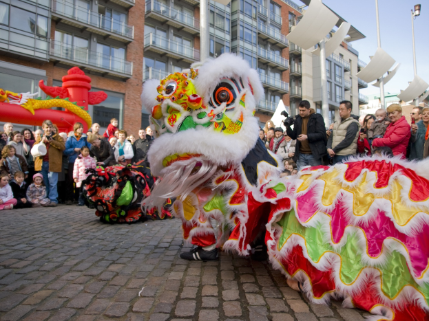Vibrantes chinesisches Neujahrsfest in Amsterdam mit einer Löwen-Tanz-Performance vor einer Zuschauermenge, darunter einige mit Kameras, vor einem Hintergrund aus Gebäuden, Laternenmasten und einem klaren blauen Himmel.