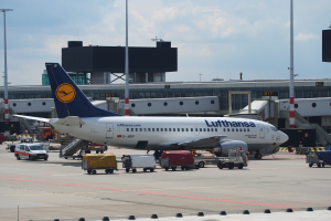 Ein Lufthansa Airbus A320-200 steht auf dem Frankfurter Flughafen geparkt, mit Fahrzeugen und Wagen auf der Straße davor, einem Gebäude, Laternenmasten und einem klaren blauen Himmel im Hintergrund.