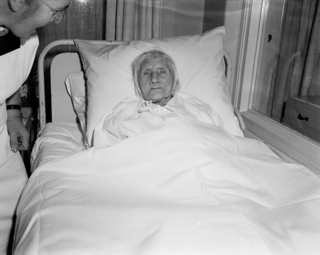 Eine ältere Frau liegt in einem Krankenhausbett mit einem weißen Laken und Kissen, während eine Krankenschwester neben ihr steht, mit einem Fenster mit Vorhängen im Hintergrund.