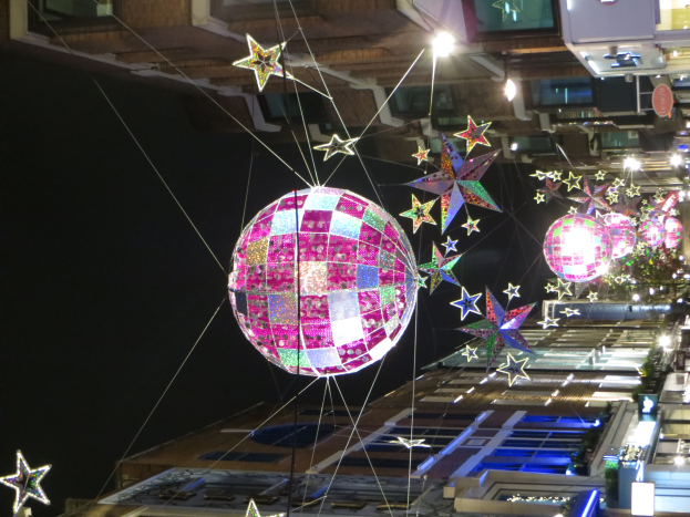 Eine nächtliche Straßenansicht einer Stadt mit zahlreichen Lichtern und Dekorationen, die einen großen Diskokugel an einem Gebäude hängen sehen lassen, der eine festliche Atmosphäre schafft.