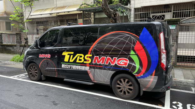 Ein schwarzer Van parkt an der Seite einer Straße neben einem Gebäude, mit Text und Zahlen darauf, und ein Auto geparkt rechts daneben, mit Gebäuden, Bäumen, Pflanzen und einem Tor im Hintergrund.