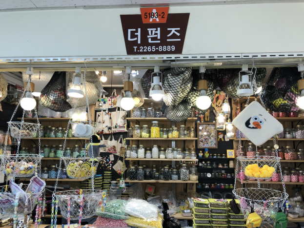 Ein Ladeneinrichtungsbild, das verschiedene Gegenstände wie Körbe, Lichter, Flaschen und Spielzeuge zeigt, die auf Regalen angeordnet sind, mit einer Tafel öber dem Eingang, die es als bestes koreanisches Geschäft in Seoul ausweist.