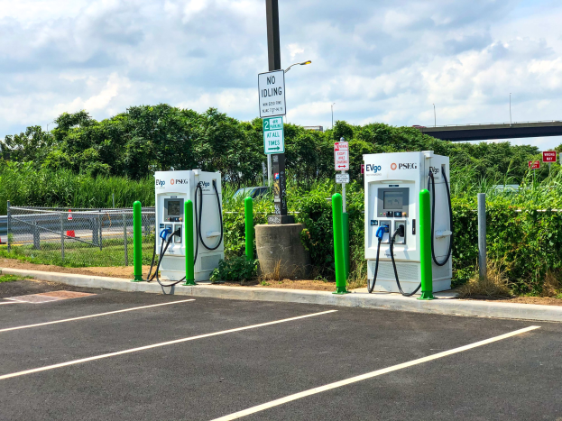 Elektrofahrzeug-Ladestation auf einem Parkplatz mit umgebender Infrastruktur und einer Brücke im Hintergrund.
