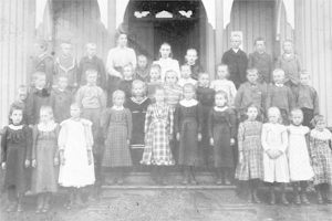 Ein Schwarz-Weiß-Foto einer Gruppe von Kindern, die vor einem Gebäude mit einer Tür und Säulen stehen, mit der Beschriftung "Erste Klasse" unten.