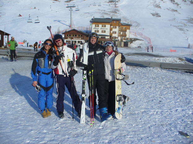 Vier Personen posieren für ein Foto mit Skating-Pads und Werkzeugen im Hintergrund, mit Gebäuden und anderen Menschen vor einer schneebedeckten Berglandschaft.
