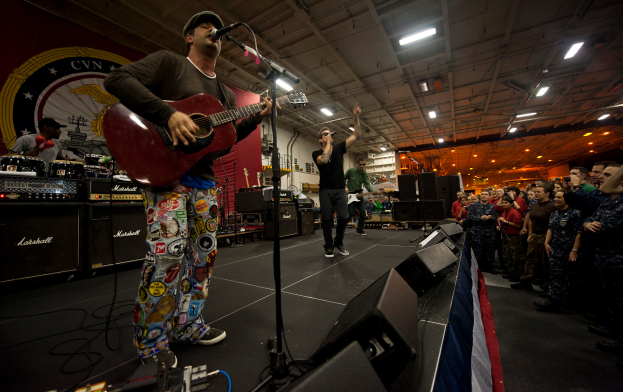 Chris Stapleton, mit einer Mütze bekleidet, spielt Gitarre auf der Bühne vor einem Publikum, umgeben von einem Mikrofon, Lautsprechern, einer Fahne und anderen Instrumenten, mit einem Banner und Lichtern im Hintergrund.